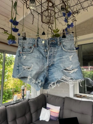 Högmidjade ljusblå jeansshorts - Trendiga ljusblå jeansshorts från Zara med slitna detaljer och fransig kant. Klassisk femficksmodell med knappgylf och bälteshällor. Perfekta för sommaren!