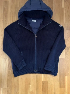 Mörkblå stickad cardigan från Moncler - Snygg mörkblå stickad cardiganjacka från Moncler med dragkedja och vadderad huva. Jackan har ribbstickade muddar, två fickor med dragkedja och Moncler-logga på ärmen. Perfekt för dig som vill ha både stil och värme i ett plagg.