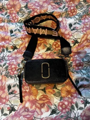 Svart axelväska Marc Jacobs - Snygg svart axelväska från Marc Jacobs i läder med bred axelrem med logga i beige. Väskan har guldfärgade metalldetaljer och en rektangulär form. Perfekt för dig som vill ha en trendig och praktisk väska med plats för det viktigaste.
