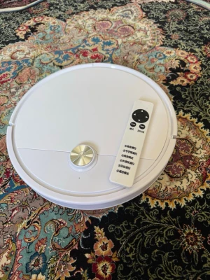 Vit robotdammsugare med fjärrkontroll - Säljer en rund, vit robotdammsugare med tillhörande fjärrkontroll. Dammsugaren har två sidoborstar undertill och en metallisk knapp på ovansidan. Perfekt för att hålla golvet rent utan ansträngning.