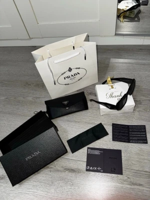 Svarta solglasögon från Prada - Snygga svarta solglasögon från Prada med rektangulär form och breda bågar. Klassisk logga på sidan och glasen är mörka. Kommer med originalfodral, putsduk och förpackning. Perfekta för dig som vill ha en stilren och lyxig accessoar.