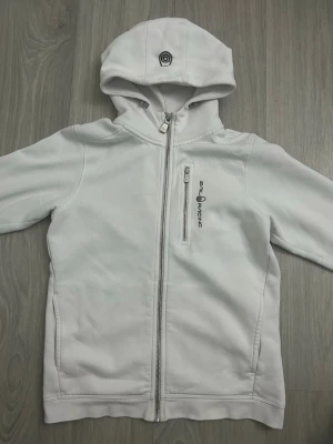Vit hoodie från Sail Racing - Snygg vit hoodie från Sail Racing med dragkedja framtill och en bröstficka med logga. Hoodien har en broderad detalj på huvan och är tillverkad i mjukt material. Perfekt för en avslappnad och sportig stil.