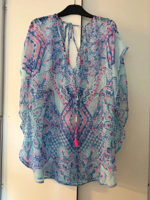 Mönstrad strandtunika i blått och rosa - Säljer en luftig strandtunika med färgglatt mönster i blått, rosa och vitt. Tunikan har v-ringning med knytband och vida ärmar. Perfekt att slänga över bikinin på stranden. Materialet är tunt och transparent, vilket gör den sval och bekväm under varma dagar. från New Yorker.