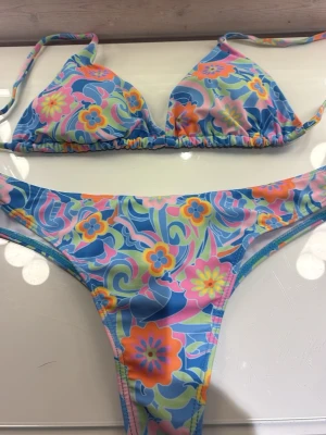Färgglad blommig bikini med knyt - Säljer en superfin trekantsbikini med blommigt mönster i blått, rosa, orange, gult och grönt. Både överdel och underdel har knytband för justerbar passform. Bikinin är helt oanvänd!Perfekt för sommaren och strandhäng!☀️