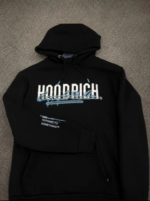Svart HOODRICH hoodie med blå detaljer - Svart hoodie från HOODRICH med stor vit och blå logga framtill och textdetalj på ärmen. Klassisk huva med dragsko och känguruficka. Snygga blå accenter på snörena och trycket ger en streetwear vibe. Perfekt för dig som gillar urban stil.