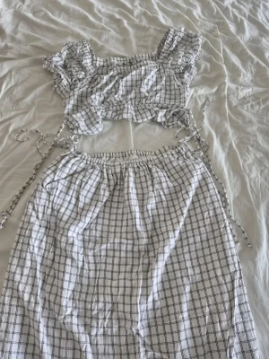 Rutigt set med topp och kjol uniqlo C S - Säljer ett vitt tvådelat set med svart rutmönster. Setet består av en kort topp med puffärmar och knytband samt en lång kjol med resår i midjan och knytband. Materialet känns lätt och luftigt, perfekt för sommaren. Bara provat. Ny utan prislapp och tvättad.