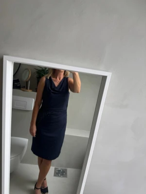 Marinblå klänning från Esprit - Säljer en marinblå klänning från Esprit med draperad halsringning och elegant fall. Klänningen är ärmlös och har en mjuk, slät polyesterkänsla som gör den bekväm och snygg. Perfekt för dig som vill ha en stilren look.