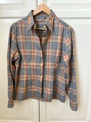 Beige rutig skjorta från Burberry - Klassisk beige rutig skjorta från Burberry med långa ärmar och knappar framtill. Skjortan har det ikoniska rutmönstret i svart, vitt och rött. Tillverkad i mjuk bomull och har en normal passform. Perfekt för dig som gillar tidlös och stilren design. Köpt secondhand så vet inte att den är helt 100