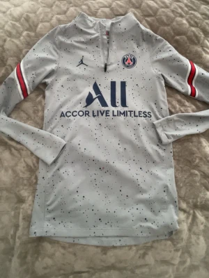 PSG x Jordan grå träningströja - Snygg grå långärmad träningströja från PSG x Jordan med blå och röda detaljer på ärmarna. Tröjan har halv dragkedja, mörkblå logga och texten 'Accor Live Limitless' framtill. Tillverkad i ett tekniskt material som passar perfekt till träning.