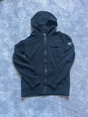 Lyle & Scott hoodie  - Svart hoodie från Lyle & Scott med dragkedja framtill, huva och ribbade muddar. Bröstficka med lock och klassisk gul logga på vänster ärm. Tillverkad i mjuk bomullsmix som är skön att ha på sig. Perfekt för en avslappnad och stilren look. Priset är inte hugget i sten!