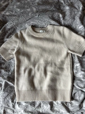 Beige stickad t-shirt med kort ärm H&M - Säljer en stilren beige stickad t-shirt med korta ärmar och bred ribbad kant vid ärmslut och nederkant. Tröjan har rund halsringning och är gjord i ett mjukt, tjockare material som känns lyxigt. Perfekt för dig som gillar minimalistisk och clean stil. Säljer tröjan då den inte kommit till användning. Skriv vid frågor!💚