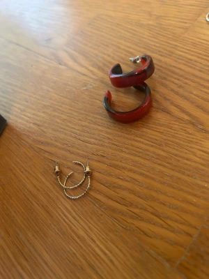 Två par örhängen i röd och silver - Säljer två par örhängen: ett par chunky röda hoops i plast med metallstift och ett par tunna silverfärgade örhängen med kedjedetaljer. Perfekta för dig som vill variera din stil med både färg och metalliska inslag.