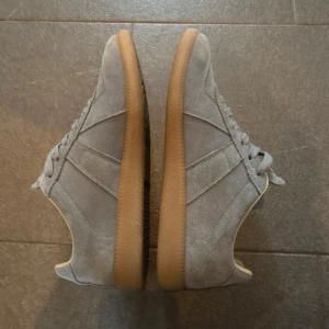 Maison Margiela | Grå suede - Snygga grå sneakers från Margiela i mocka med klassisk design och gummisula i beige. Skorna har lågt skaft, matchande grå skosnören och diskreta sömdetaljer på sidan. Perfekta för en clean och stilren look.