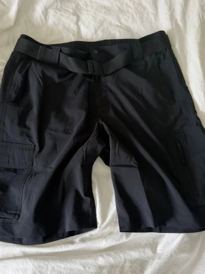 Svarta shorts från Columbia med bälte - Svarta shorts från Columbia med praktiskt bälte och flera fickor, inklusive en med dragkedja. Perfekta för sommarens äventyr och outdoor-aktiviteter. Tillverkade i ett lätt syntetmaterial som andas och torkar snabbt.