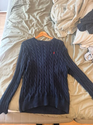 Mörkblå kabelstickad tröja Polo Ralph Lauren - Snygg mörkblå kabelstickad tröja från Polo Ralph Lauren med rund halsringning och röd broderad logga på bröstet. Tröjan har långa ärmar och ribbade muddar vid ärmslut och nederkant. Perfekt för dig som gillar klassisk och tidlös stil.