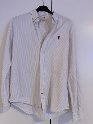 Vit randig skjorta från U.S. Polo Assn. - Klassisk vit skjorta med tunna ljusgrå ränder från U.S. Polo Assn. Skjortan har lång ärm, knappar framtill och en liten broderad logga på bröstet. Tillverkad i bomull och har en tidlös, clean look som passar till mycket.