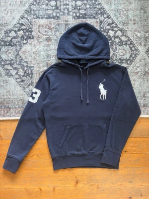 Marinblå Big Pony Polo Ralph Lauren Hoodie - Storlek S - Hej! 👋 Säljer en riktigt fräsch marinblå Polo Ralph Lauren ”Big Pony” hoodie i storlek S. Har den stora broderade vita loggan på bröstet och den klassiska 3-an på höger ärm. 🐎✨ Tröjan är i mycket gott skick, välvårdad och helt utan hål eller fläckar. Garanterat 100% äkta med originaltvättlappar kvar (saknar QR-kod pga årsmodell). Ett tidlöst premium-basplagg med helt rätt sportig look som passar till allt! Fraktar snabbt! 🚛📦💬 
