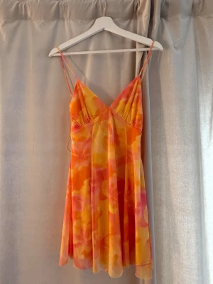 Färgglad kort klänning med snörning - Superfin kort klänning från Gina Tricot med tunna axelband och snörning i ryggen. Klänningen har ett färgstarkt blommigt mönster i orange, gul och rosa toner, samt v-ringad hals. Perfekt för sommarens alla tillfällen!