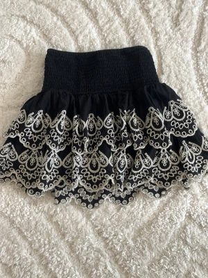 Svart kortkjol med broderi från Lindex - Supersöt svart kortkjol från Lindex med bred resår i midjan och vita broderade detaljer i ett vågigt mönster. Kjolen har volangkant och är perfekt för dig som gillar en lekfull och trendig stil.