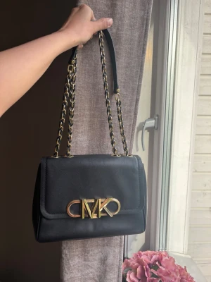 Svart Michael Kors handväska i skinn - Snygg svart handväska från Michael Kors i slätt skinn med guldfärgad kedjerem och stort MK-logo framtill. Väskan har en klassisk rektangulär form och detaljer i metall som ger en lyxig känsla. Perfekt accessoar för att lyfta din outfit.