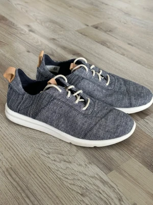 Gråmelerade sneakers med vita sulor från Toms - Snygga sneakers i gråmelerat tyg med vita sulor och beige detaljer på häl och plös. Skorna har vita snören och en låg profil som ger en clean och modern look. Perfekta för dig som gillar stilrena och bekväma skor till vardags.