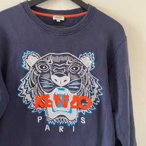 Blå Kenzo sweatshirt med tigerbrodyr - Säljer en marinblå sweatshirt från Kenzo med ikonisk tigerbrodyr och orange logga framtill. Tröjan har rund hals, långa ärmar och är gjord i mjuk bomull. Perfekt statement piece för dig som vill sticka ut med cool design och snygga detaljer. Nypris: 3999kr