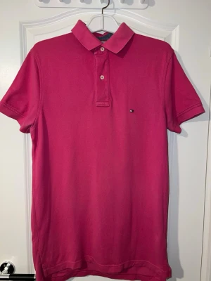 Rosa pikétröja från Tommy Hilfiger - Snygg rosa pikétröja från Tommy Hilfiger med klassisk krage och två knappar framtill. Tröjan har korta ärmar och den ikoniska lilla loggan broderad på bröstet. Perfekt för en clean och stilren look. Finns att hämta i Södertälje.