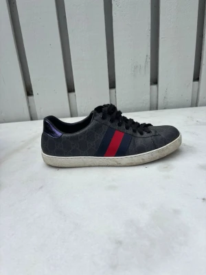 Gucci Ace GG Supreme - Gucci sneakers i svart med monogrammönster, röda och blå ränder på sidorna samt metallicröd och metallicblå häl. Klassisk låg modell med vita sulor och svarta skosnören. Snygg och tidlös design som sticker ut. Boxen har varit i solen så den är lite färgblek. Pris kan diskuteras. Kan självklart tvära skorna innan 