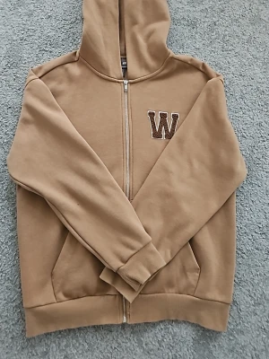 Beige hoodie med dragkedja från H&M - Säljer en beige hoodie från H&M i relaxed fit med dragkedja framtill och broderad W på bröstet. Hoodien har huva, ribbade muddar och fickor på sidorna. Perfekt för en chill och avslappnad stil. Aldrig använd.