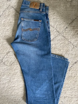 Blå raka jeans från Nudie Jeans - Säljer ett par klassiska blå raka jeans från Nudie Jeans med ikonisk bakficka och kontrastsömmar. Jeansen har normal midja och är tillverkade i slitstark denim. Perfekta för en avslappnad och stilren look.