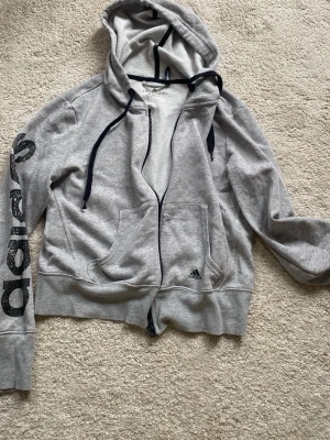 Grå zip-hoodie från Adidas - Grå hoodie från Adidas med dragkedja, svart snörning i huvan och stora fickor framtill. Adidas-logga på bröstet och tryck längs ena ärmen. Tillverkad i mjuk climalite-bomull som andas. Perfekt för en sportig och avslappnad stil.