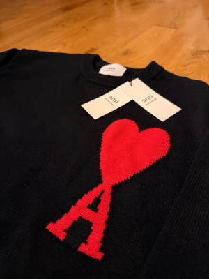 Svart stickad tröja från Ami Paris - Svart stickad tröja från Ami Paris med rund halsringning och ribbade muddar. På bröstet finns ett stort rött hjärta och bokstaven 'A' i samma färg. Tröjan har en klassisk passform och är perfekt för dig som gillar stilrena detaljer.