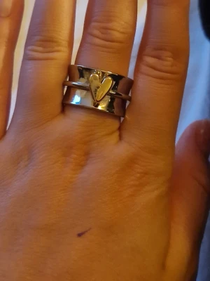 Silverfärgad ring med guldfärgat hjärta - Stilren och bred silverfärgad ring med ett upphöjt guldfärgat hjärta i mitten. Ringen har en modern och chunky design med slät yta och passar perfekt för dig som gillar att sticka ut med dina accessoarer. Materialet ser ut att vara metall i både silver- och guldfärg. Aldrig använd helt ny. Pris kan diskuteras.