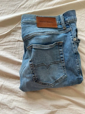 Ljusblå jeans från Diesel - Säljer ett par ljusblå jeans från Diesel med klassisk femficksdesign och skinnpatch med logga bak i midjan. Jeansen har raka ben och är tillverkade i mjukt denimtyg. Perfekta för en avslappnad och trendig look.