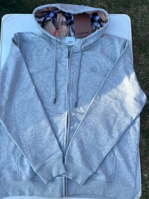 Grå zip hoodie från Burberry - Snygg grå zip hoodie från Burberry med klassiskt rutigt foder i huvan och broderad logga på bröstet. Hoodien har dragsko i huvan, fickor fram och ribbade muddar. Perfekt för dig som vill ha en stilren och exklusiv look.