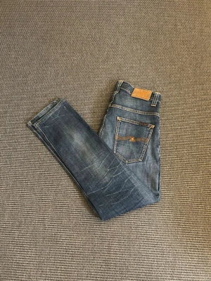 Nudie jeans - Säljer ett par klassiska blå jeans från Nudie Jeans med snygga slitningar och kontrastsömmar. Jeansen har en rak passform och detaljerad bakficka med Nudie-loggan. Tillverkade i denim med cool vintagekänsla.