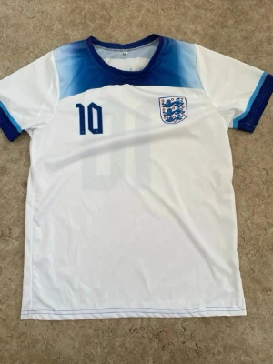 England fotbollströja Bellingham #10 - Säljer en vit England fotbollströja med blå detaljer, nummer 10 och Bellingham på ryggen. Tröjan har korta ärmar, rund hals och Englands landslagsmärke på bröstet. Perfekt för fotbollsfans som vill visa sitt stöd under matcherna.