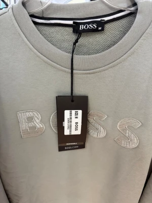 Ljusgrå sweatshirt från BOSS - Snygg ljusgrå sweatshirt från BOSS med broderad logga framtill. Tröjan har rund halsringning, ribbade muddar vid ärmslut och nederkant samt en clean och stilren look. Perfekt för dig som gillar minimalistisk design och premiumkänsla.