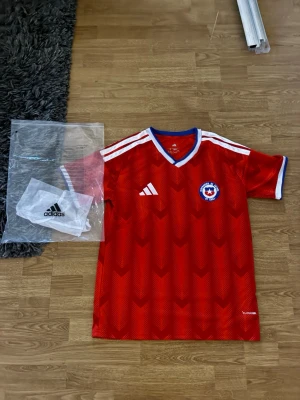 Chile Hemmaställ - Snygg röd fotbollströja från Adidas med Chile landslagsmärke på bröstet. Tröjan har vita detaljer, blå och vit V-ringad krage samt klassiska Adidas-ränder på axlarna. Tillverkad i lätt och ventilerande material, perfekt för träning eller match.