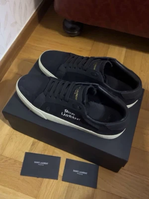  Saint Laurent sneakers svarta - Svarta sneakers från Saint Laurent med vit sula och handskriven logga på sidan. Skorna har klassisk låg modell, svarta skosnören och detaljer i guld på plösen. Tillverkade i canvas och läder för en stilren och lyxig känsla. Box och distraherad ingår!