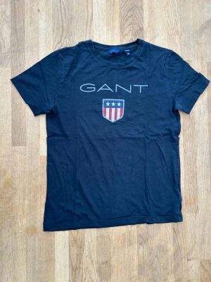 Svart GANT t-shirt med logga - T-shirt från GANT med klassisk logga och sköld med stjärnor och ränder på bröstet. T-shirten har rund halsringning och korta ärmar. Tillverkad i mjuk bomull, perfekt för en avslappnad stil.