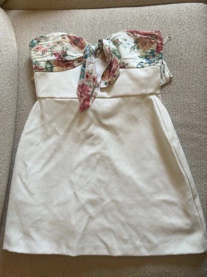 Vit axelbandslös klänning med blommor - Jätte populär och  söt axelbandslös kortklänning från zara, med vit kjol och blommigt mönster upptill i rosa, blått och grönt. Överdelen har knyt fram och ger en romantisk vibe. Helt slutsåld på hemsidan!😊 Perfekt för sommarens alla tillfällen. Skriv privat vid frågor och funderingar!
