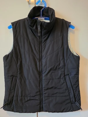 Columbia Puffer Vest Black Women's L | Vadderad Väst Outdoor - Snygg och varm Columbia-väst i svart med diskret broderad logga. Perfekt för höst, vår och lager-på-lager under kallare dagar. Dragkedjefickor och hög krage. Fint begagnat skick.  Storlek: Women's L Skick: Mycket fint skick Märke: Columbia Sportswear Company  Skickas snabbt! Oftast samma dag!  Marttila Vintage
