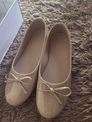 Beige ballerinaskor med rosett - Snygga beige ballerinaskor från Kelly med glansig finish och söt rosett framtill. Skorna har rund tå och klassisk design, perfekta för en stilren look. Tillverkade i syntetmaterial med mjuk innersula för extra komfort.