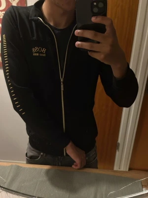 Svart ziptröja från Hugo Boss - Svart ziptröja från Hugo Boss med guldfärgad dragkedja och logga på bröstet. Tröjan har gula detaljer längs ärmarna och en klassisk krage. 