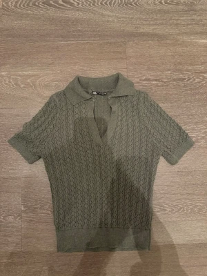 Olivgrön stickad topp från Zara - Snygg olivgrön stickad topp från Zara med korta ärmar och krage. Djup v-ringning framtill och diskret mönster i stickningen ger en trendig look. Perfekt till jeans eller kjol för en cool vardagsstil.