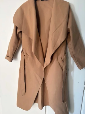 Camel kappa med oversized krage Boohoo - Stilren camel-färgad kappa från Boohoo med oversized krage och öppen front. Kappan har långa ärmar, två snedställda fickor och ett mjukt tyg som ger en avslappnad look. Perfekt för lager-på-lager och trendig streetstyle.
