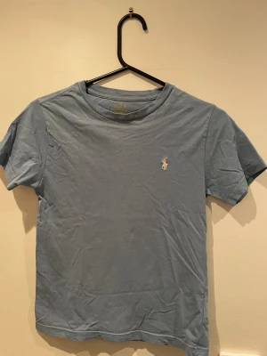 Blå t-shirt från Polo Ralph Lauren - Säljer en klassisk blå t-shirt från Polo Ralph Lauren med den ikoniska loggan broderad i vitt på bröstet. T-shirten har rund halsringning och korta ärmar. Perfekt basic-plagg till garderoben.