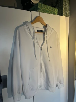 Vit zip hoodie från Polo Ralph Lauren - Snygg vit hoodie med dragkedja från Polo Ralph Lauren. Klassisk design med huva och dragsnören, två stora fickor framtill och den ikoniska lilla loggan broderad på bröstet. Tillverkad i mjukt bomullsmaterial som är skönt att ha på sig.