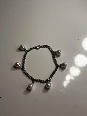 Silverfärgat armband med pärldroppar - Snyggt armband i silverfärgad metall med sex vita droppformade pärlor som hänger längs kedjan. Perfekt för dig som gillar en stilren men ändå unik accessoar. Lätt att matcha med andra smycken och ger en cool touch till din outfit.